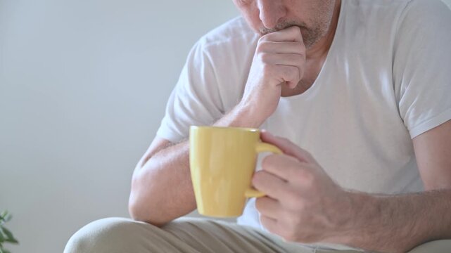 Portrait of tired mature man sitting in living room in the morning, holding yellow coffee cup and rethinking with weary expression. 4K.