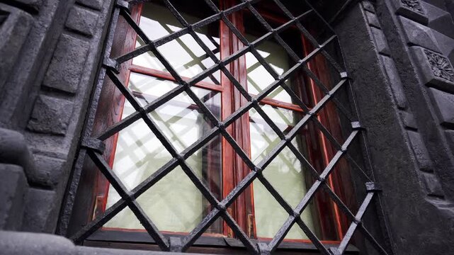 Close up view dark ornate iron grate covering wooden window of old stone building facade. Architectural detail includes carved stone elements and reflections glass.