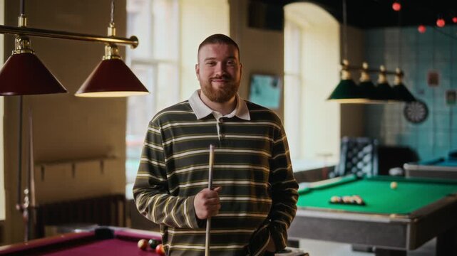 Dolly in portrait shot of bearded young adult man smiling at camera while posing with cue at billiard hall, slow motion