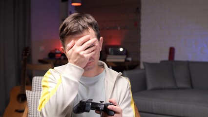 Unhappy gamer man experiencing disappointment and frustration while playing a video game at home, holding a controller and covering face with his hand after a gaming loss © Aliaksandr