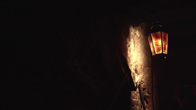 Warm glow of vintage lantern against rough texture of old stone wall in deep darkness, highlighting architectural detail and creating sense of history and mystery.