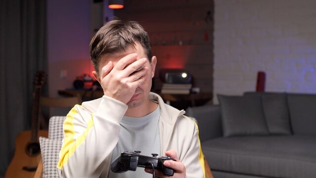 Unhappy gamer man experiencing disappointment and frustration while playing a video game at home, holding a controller and covering face with his hand after a gaming loss