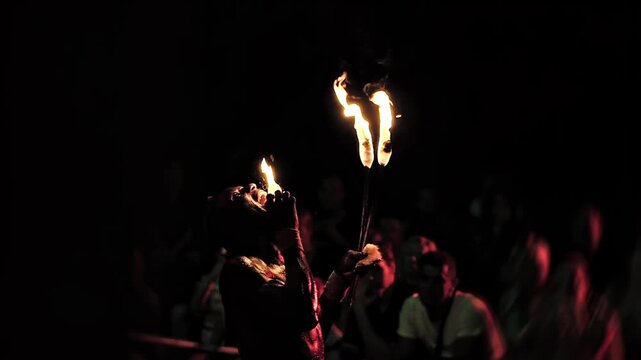 Closeup of dark-skinned performer demonstrating fire stunt, placing glowing ember from torch onto tongue, smoke rises against dark background of night, audience watches intriguing and dangerous specta