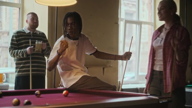 Young adult woman aiming at pool ball with cue then making successful shot cheered on by friends while hanging out at bar over beers