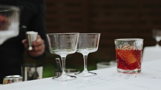 Two empty glasses on the bar counter in an outdoor cafe. Bartender preparing to make a cocktail, close-up of Negroni, mixologist measuring bitters with a jigger over a white menu.