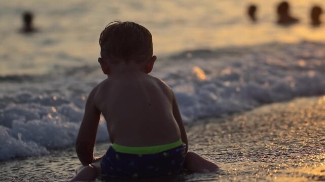 Boy, rear view, observes gentle ocean waves on shore at sunset. Warm light reflects on wet sand and water surface. Serene moment of leisure and relaxation. Vacation holiday leave sea. Slow motion