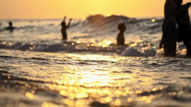 Warm golden sun reflection creates sparkling bokeh effect on ocean water surface and wet sand. Small waves break near shore. Blurry figures of people enjoy recreational swim. Vacation holiday leave se