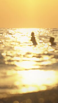 Abstract view of unrecognizable individuals enjoying swim in sea at dusk. Sun reflection shimmers on water, creating beautiful bokeh lights. Serene coastal evening atmosphere. Vertical shot. Summer va