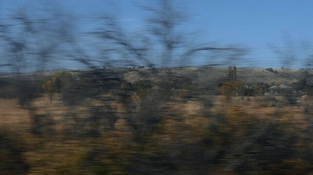 Expansive rural landscape captured with motion blur foreground of dry fields and blurred trees, revealing distant hills, power lines, and scattered houses under bright clear blue sky, suggesting movem
