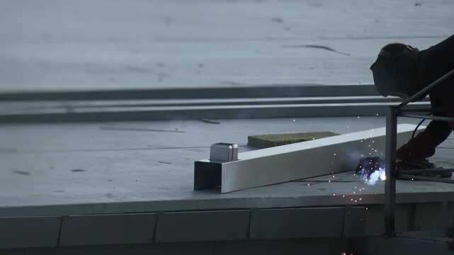 Side view of worker wearing protective helmet and gloves, expertly joining metal beams on flat gray rooftop. Bright sparks illuminate area, highlighting craftsmanship of skilled tradesperson. Hard wor