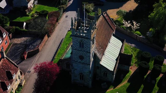 A close-up, high-angle aerial view of a historic stone church situated in a quiet English village. The shot heavily features the intricate architecture of the square church tower