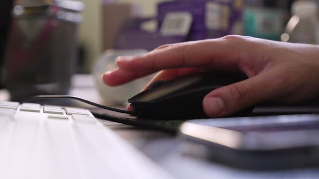 a person's hand using a black computer mouse on a desk with a white keyboard and smartphone. Shallow depth of field in a home office during the day.