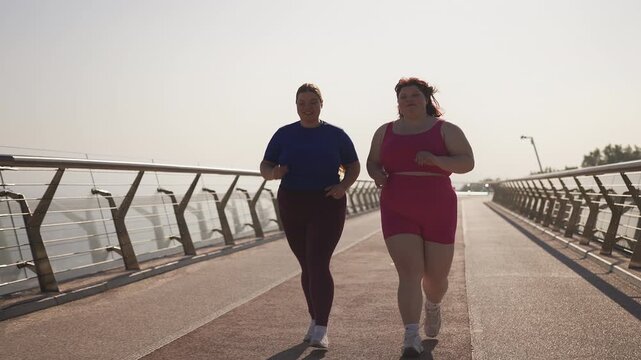 Two overweight female friends in activewear jogging across a bridge, weight loss