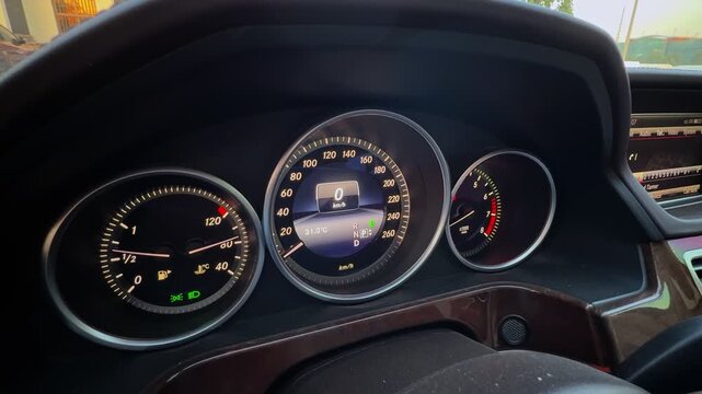 a high-end car instrument cluster. Digital speedometer shows zero km h during the day with illuminated gauges and polished wood trim accents in a modern vehicle cabin.