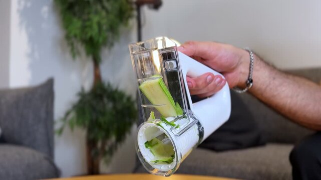 hands using a white handheld electric slicer to cut zucchini into a bowl. Bright indoor setting during the day, focusing on healthy meal preparation and kitchen gadgets.