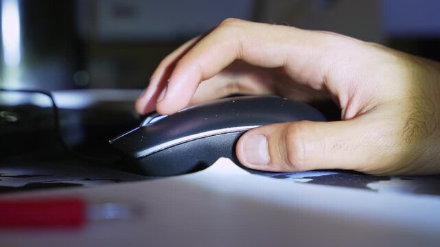 a person's hand using a computer mouse at a desk during the night. Low light creates a cinematic workspace atmosphere with blurred office supplies in the foreground.