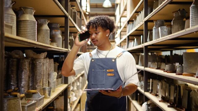 Professional African American warehouse worker in a denim apron talks on a smartphone while checking stock inventory with a clipboard amidst shelves of ceramic pottery in a storage facility.