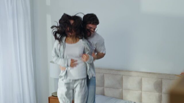 Happy Caucasian couple wife husband man woman jump on bed bedroom apartment fun boyfriend girlfriend jumping holding hands enjoyment laugh female male active bounce pampering move energy home together