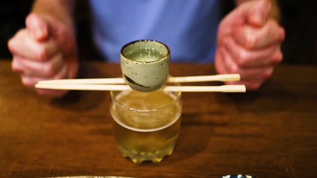 A man creates a sake bomb in slow motion by dropping a shot glass into a glass of beer resulting in a dramatic splash and foam.