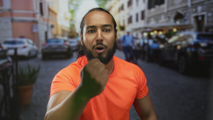 Fototapeta premium Man in orange tshirt with clenched fist shouting on a crowded city street near parked cars and sidewalk cafes; anger defiance.
