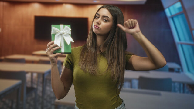 Woman holding green gift box with white bow, thumb down gesture and pursed lips in a classroom building; disappointment rejection.