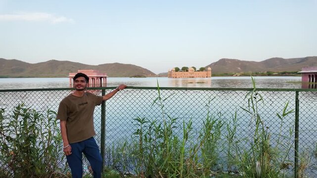 A young man leans on a fence with a view of Jal Mahal palace.