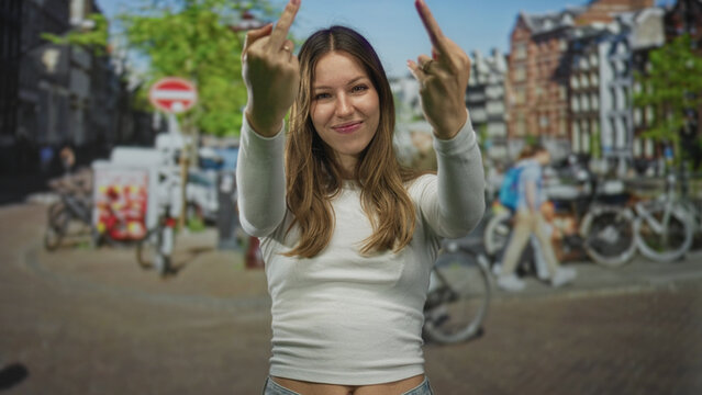 Woman showing both middle fingers toward camera on a busy city street with hands extended and a slight smile, casual white top; defiance attitude.