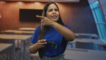 Young hispanic woman points finger while holding a gamepad in a studio, gesturing toward an offscreen display  determined concentration. © Krakenimages.com