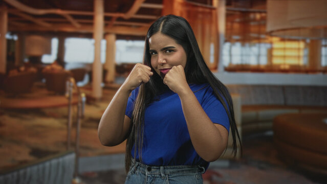 Young woman with fists raised in a boxing pose, punching motion and guarded stance inside a building lounge; confidence strength playfulness.