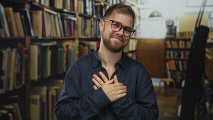 Man with hand on chest waving amid bookshelves in a building library aisle, wearing glasses and a navy shirt, smiling  gratitude warmth. © Krakenimages.com