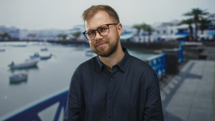Fototapeta premium Man smiling with glasses and visible face on a seaside street promenade, hands relaxed; serenity confidence.