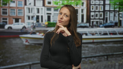 Fototapeta premium Woman with hand on chin and folded arm near a canal street in amsterdam with canal boat and narrow historic buildings visible; confidence.