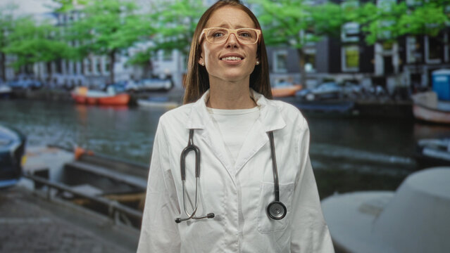 Young woman doctor in white coat with stethoscope raising hands on canal street in amsterdam by moored boats and waterway; medical care concern.