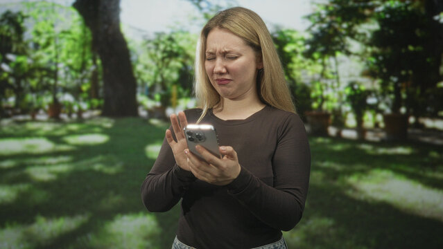 Woman holding smartphone with a grimace and recoiling hand in a sunlit garden, pulling the device away while looking disgusted; disgust repulsion surprise.