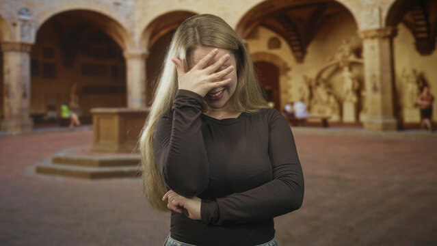 Woman blonde covers face with hand and presses forehead, standing in a historic courtyard inside a building with arches and statues; embarrassment introspection.