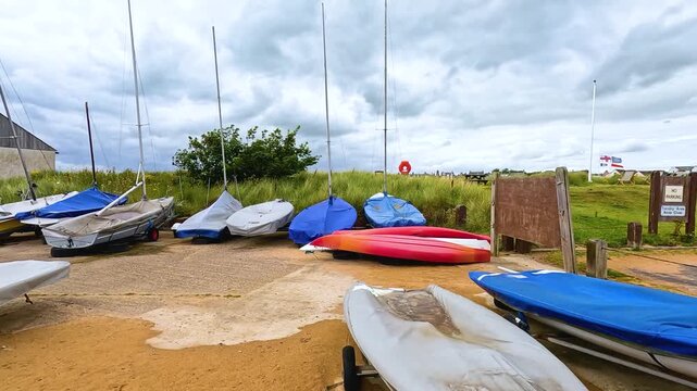 Panning shot of various sailboats and kayaks parked on beach boat yard under cloudy sky.