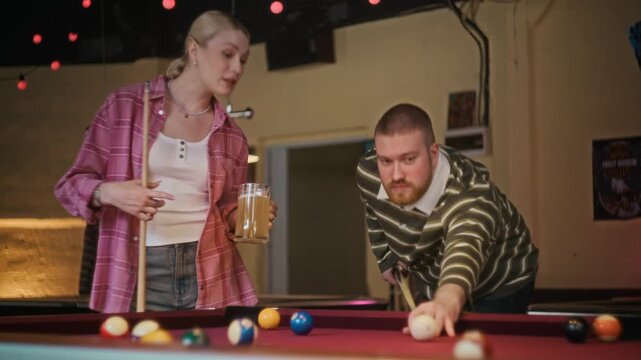 Medium full shot of young adult woman holding mug of beer guiding male friend through billiard game strategy while pointing at balls on red felt table at pool club then man striking strong shot