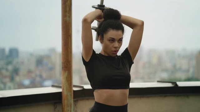 Strong Black female doing overhead triceps extension with dumbbell on rooftop