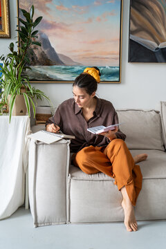 Relaxed woman painter enjoying art hobby therapy, drawing pictures with watercolor comfortably sitting on couch at home loft workshop, tranquil female artisan painting in album using aquarelle palette