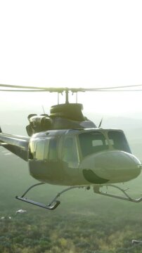 A helicopter hovers in the sky, flying over a vast landscape with hills and valleys. The sun sets in the background, casting a warm light on the scene.
