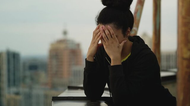 Upset Black woman covering face with hands, thinking about problems on rooftop