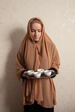 young muslim woman in beige hijab holds a copper tray with finjan cups of arabic coffee, ideal for ramadan hosting, qahwa ritual