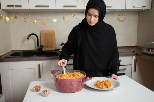 woman serves freshly cooked orange rice from a large pot onto a white plate in a bright home kitchen, illustrating meal serving, food preparation and homemade festive cuisine concepts