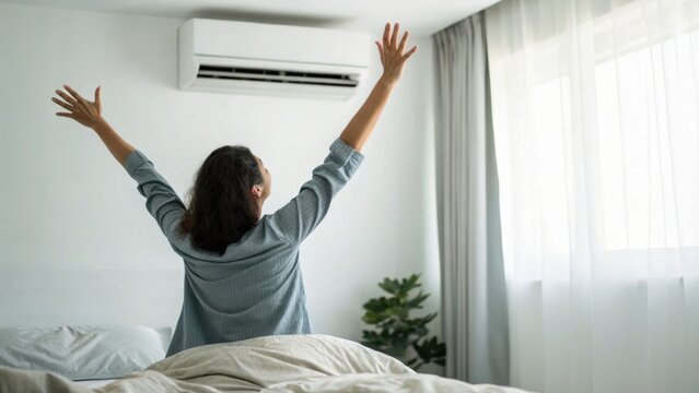 A person stretching their arms in bed near a window with natural light and an air conditioner mounted on the wall.