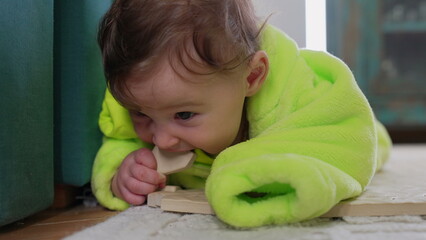 Baby lying on floor in bright green fleece outfit, exploring object by mouthing during sensory development stage at home © Marco
