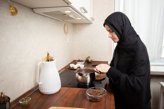 muslim woman in black hijab preparing arabic coffee on stovetop beside a white dallah, coffee beans and cardamom in a bright home kitchen during ramadan