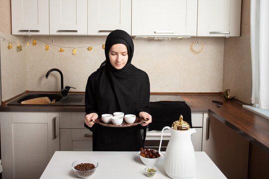 muslim woman in black hijab holding a copper tray with finjan cups prepared for arabic coffee service in a clean home kitchen with dates and beans nearby