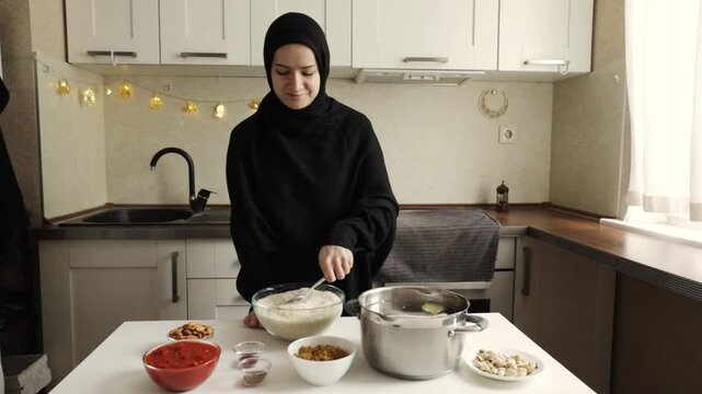 smiling muslim woman in black hijab stirring rice in large bowl beside cooking pot and ingredients during home meal preparation for ramadan gathering