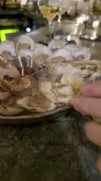 Person picking up shucked oyster from platter as empty shells are flipped, white wine glasses in background, vertical