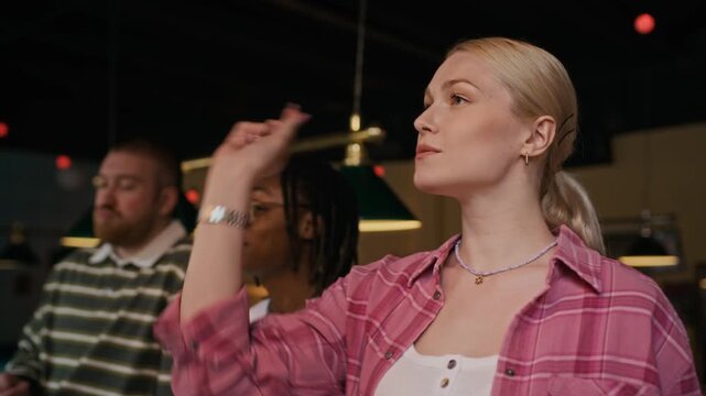 Medium shot of young adult woman throwing darts at dartboard during competition with cheerful friends at entertainment venue